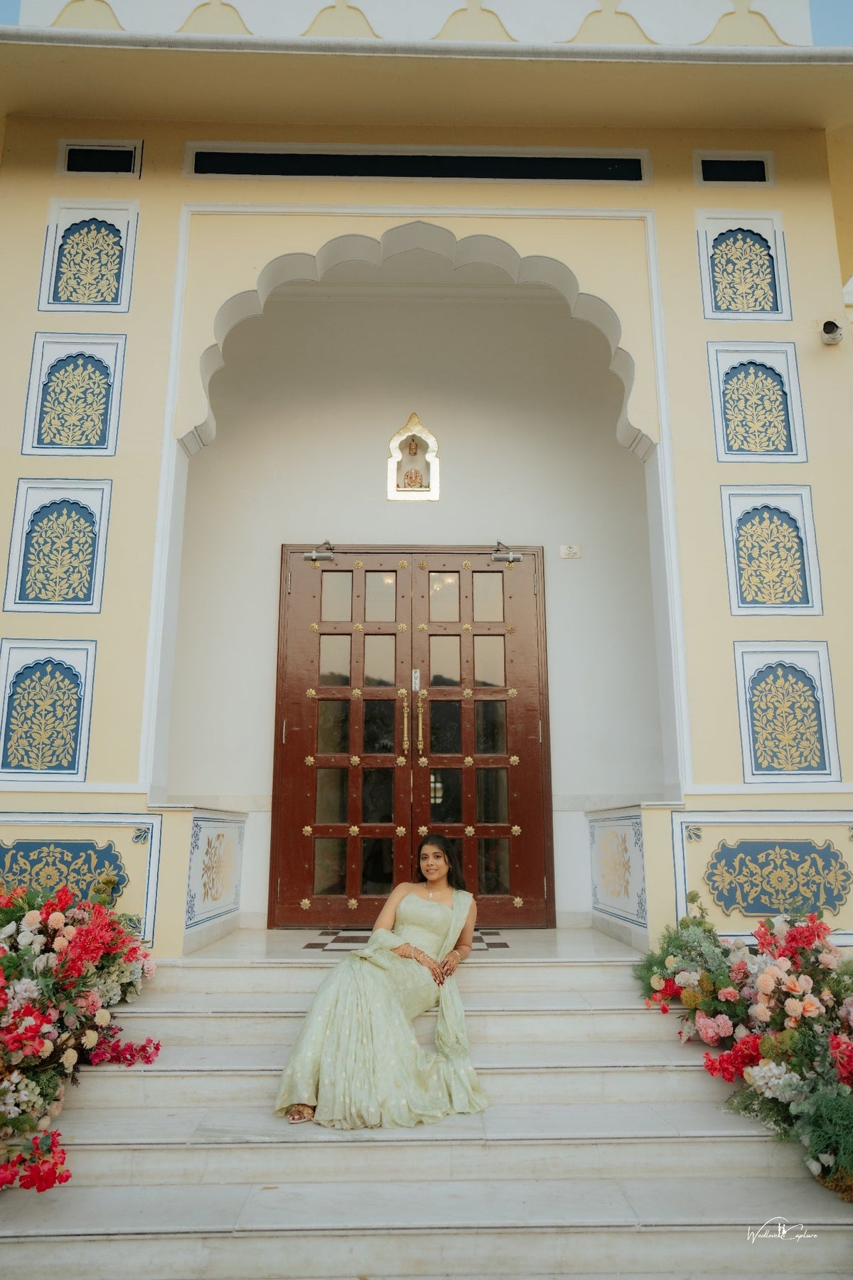 Jinali Shah in a sage green jacquard corset with drape skirt and stole, seated on palace steps before an ornate doorway