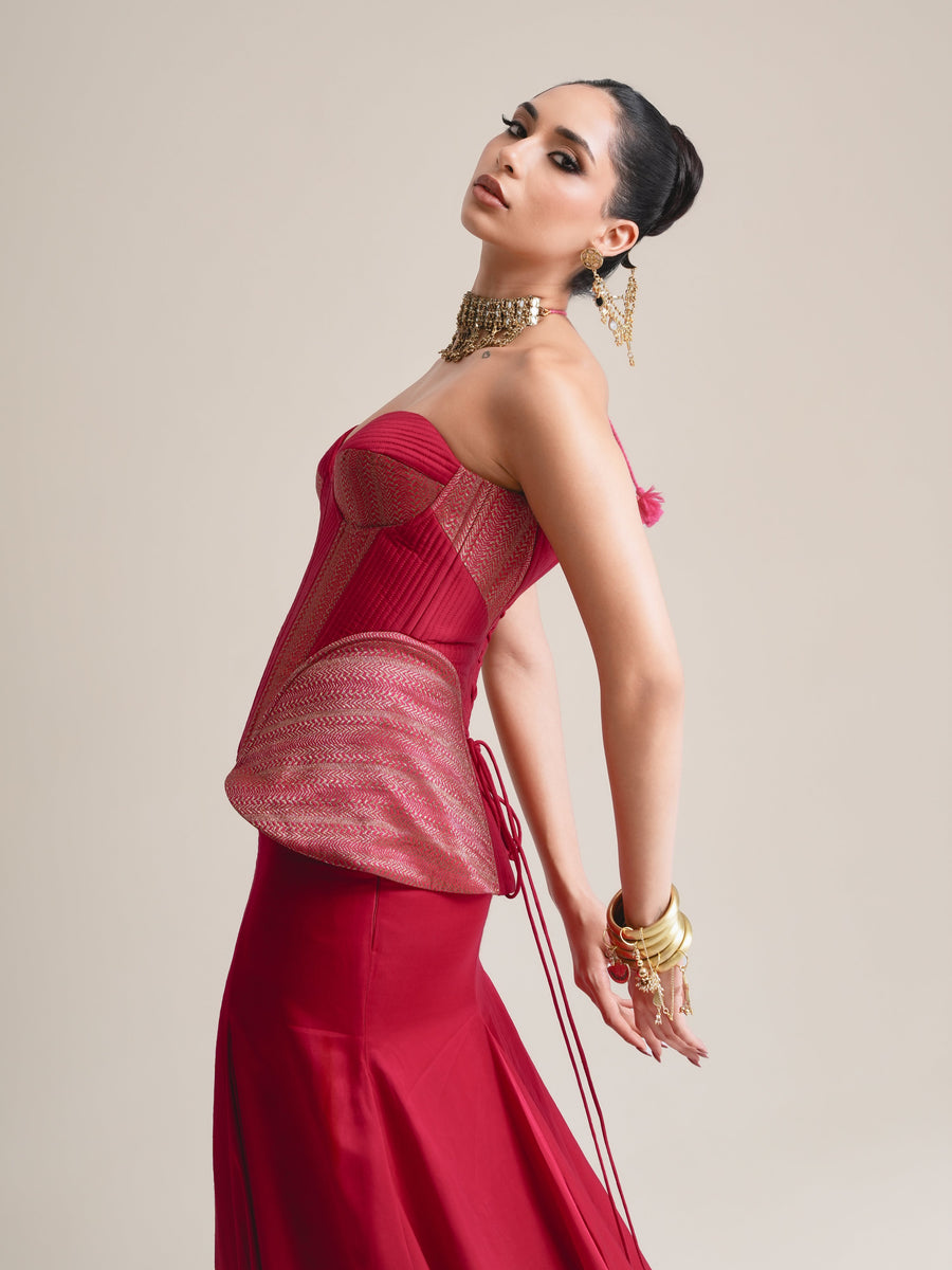 Three-quarter view of the crimson corset with rose-pink panelled peplum, head tilted back, wrists layered with gold bangles