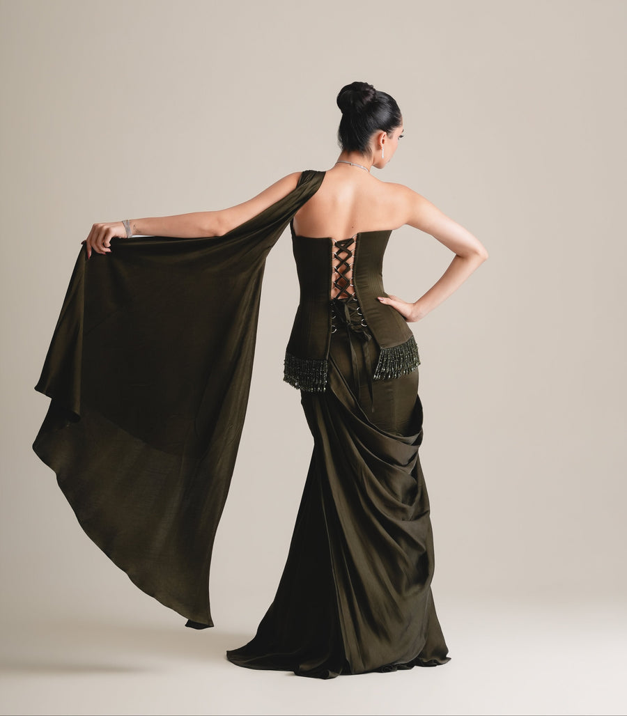 Back view of the olive-black corset with lace-up back and beaded hip fringe, the drape held out wide to one side, over a draped skirt, full length