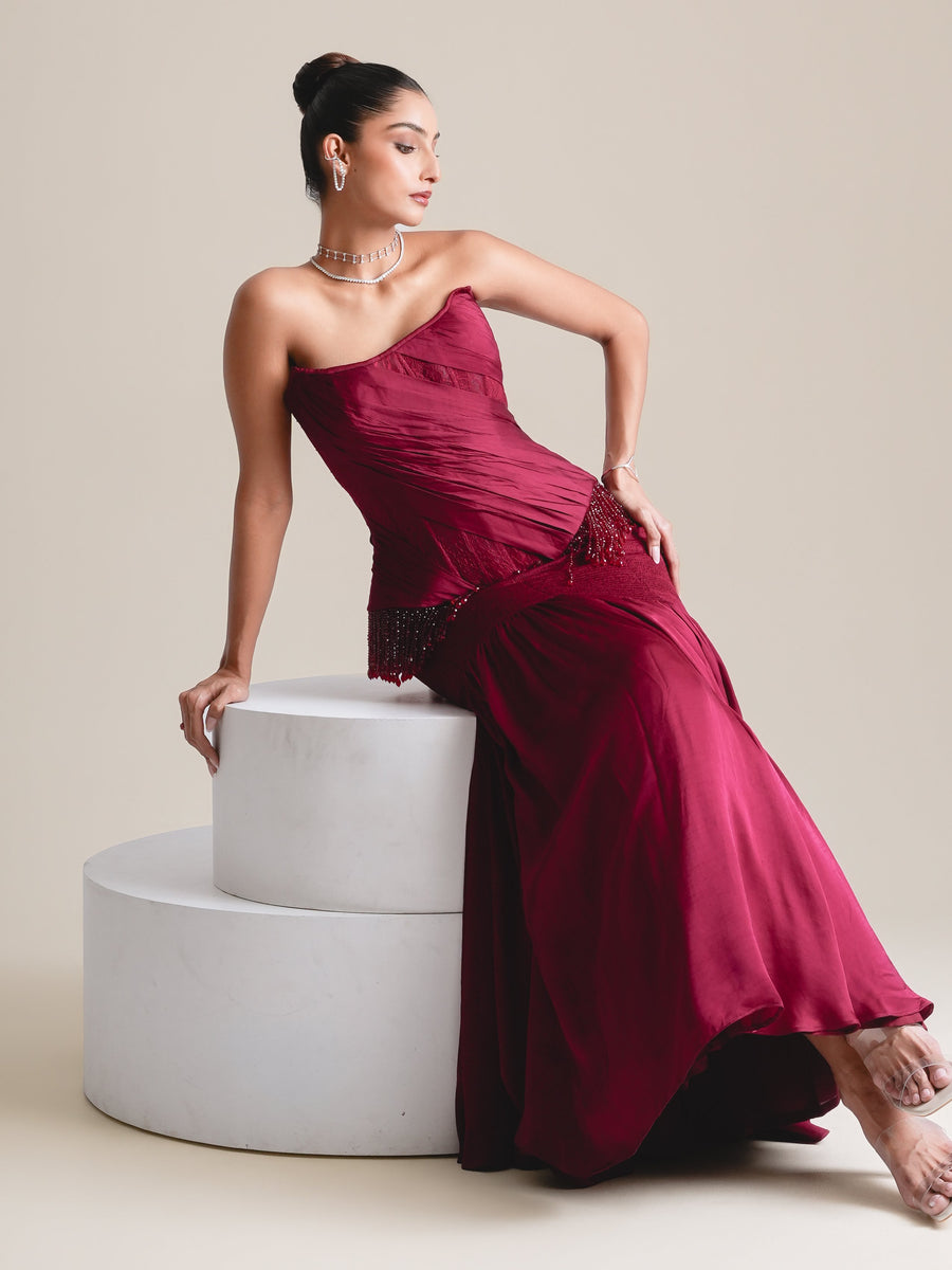 Wine-red strapless corset and long skirt, full-length view seated on white plinths, one hand on the plinth