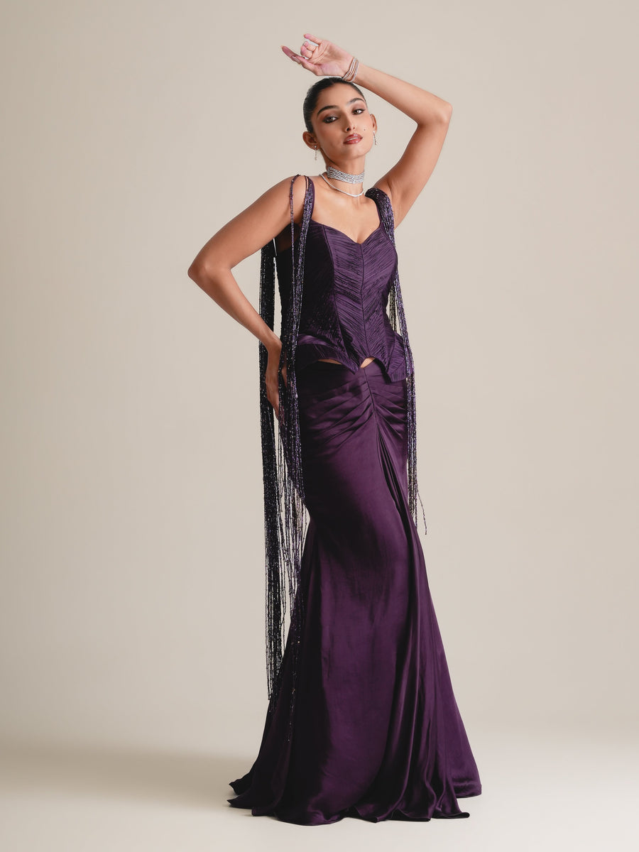 Deep-purple structured corset with long beaded tassel fringe falling from the straps over a fishcut skirt, full-length front view, one hand raised to the head