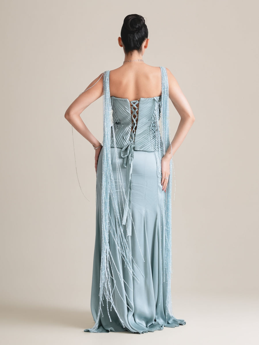 Back view of the ice-blue pleated corset with lace-up back and long fringe drapes over a floor-length skirt, full length, hands on hips
