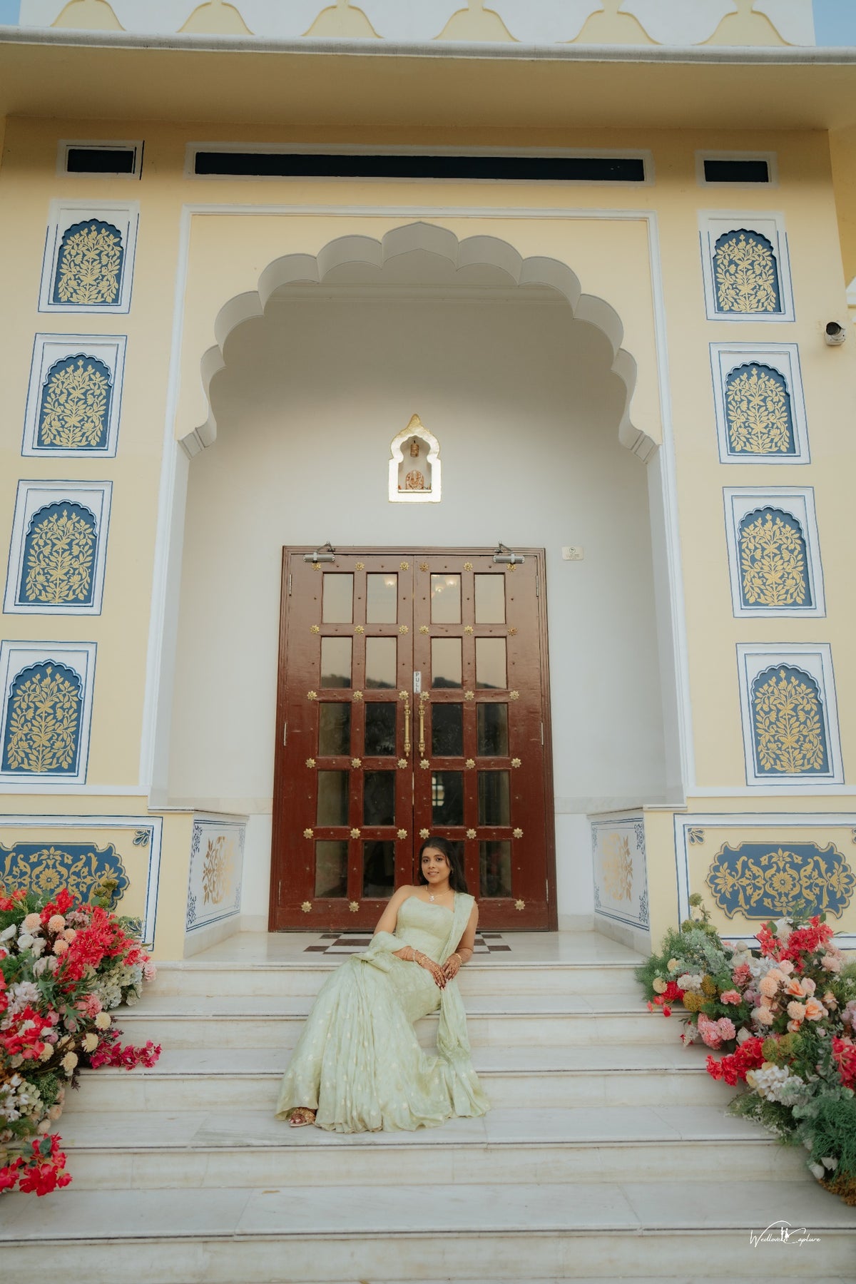 Jinali Shah in a sage green jacquard corset with drape skirt and stole, seated on palace steps before an ornate doorway