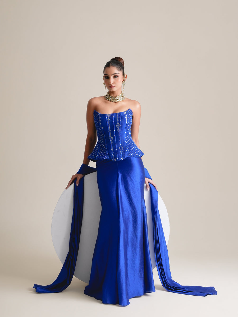 Electric blue embroidered corset and satin skirt with stole, full-length view seated against a white plinth