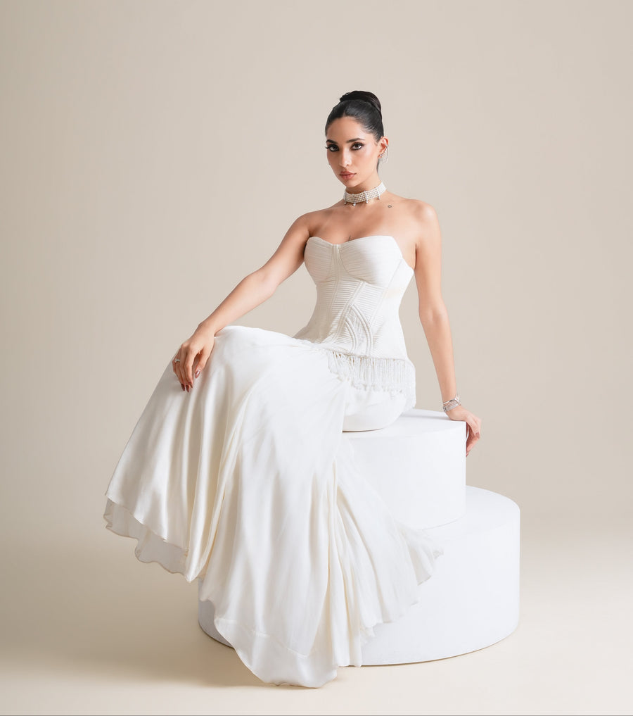 Ivory corset with fringed peplum and flowing drape skirt, full-length view seated on a white plinth